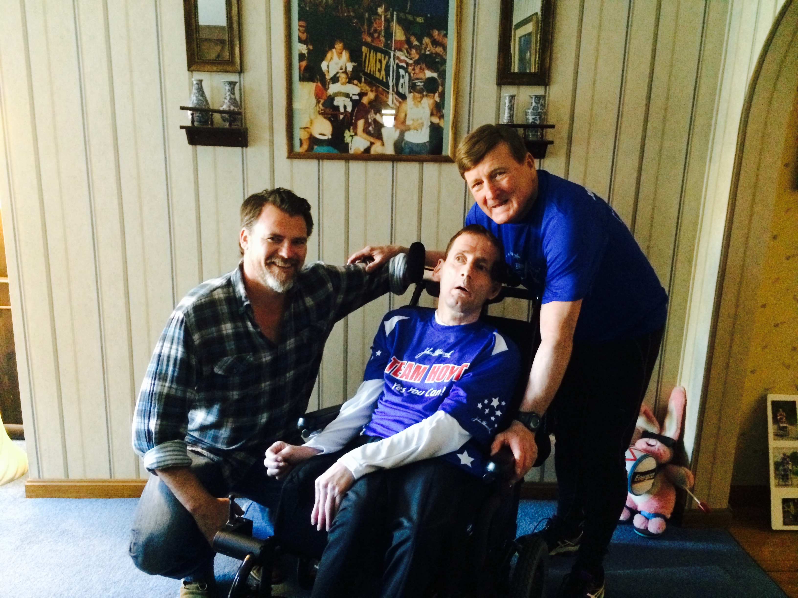 Eric kneeling beside Rick Hoyt in his Team Hoyt jersey, with Dick Hoyt leaning in behind them at the family home