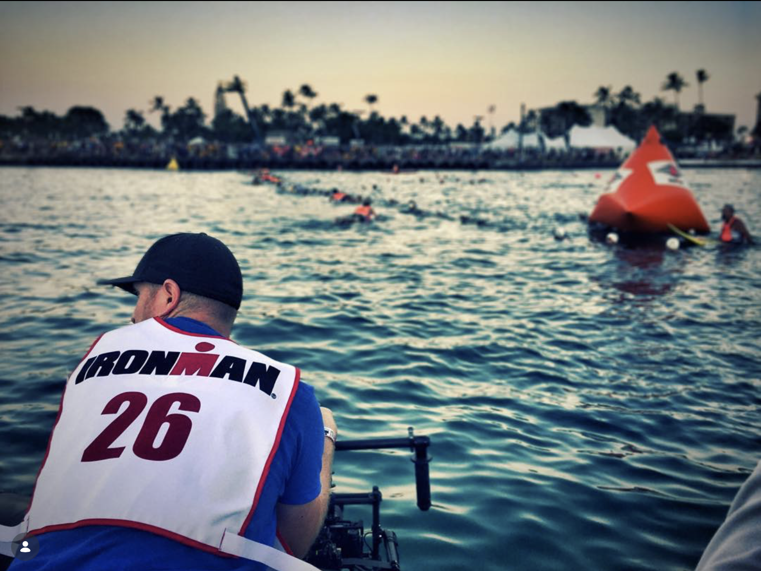 A camera operator in an IRONMAN vest shooting the swim start from a boat at sunrise