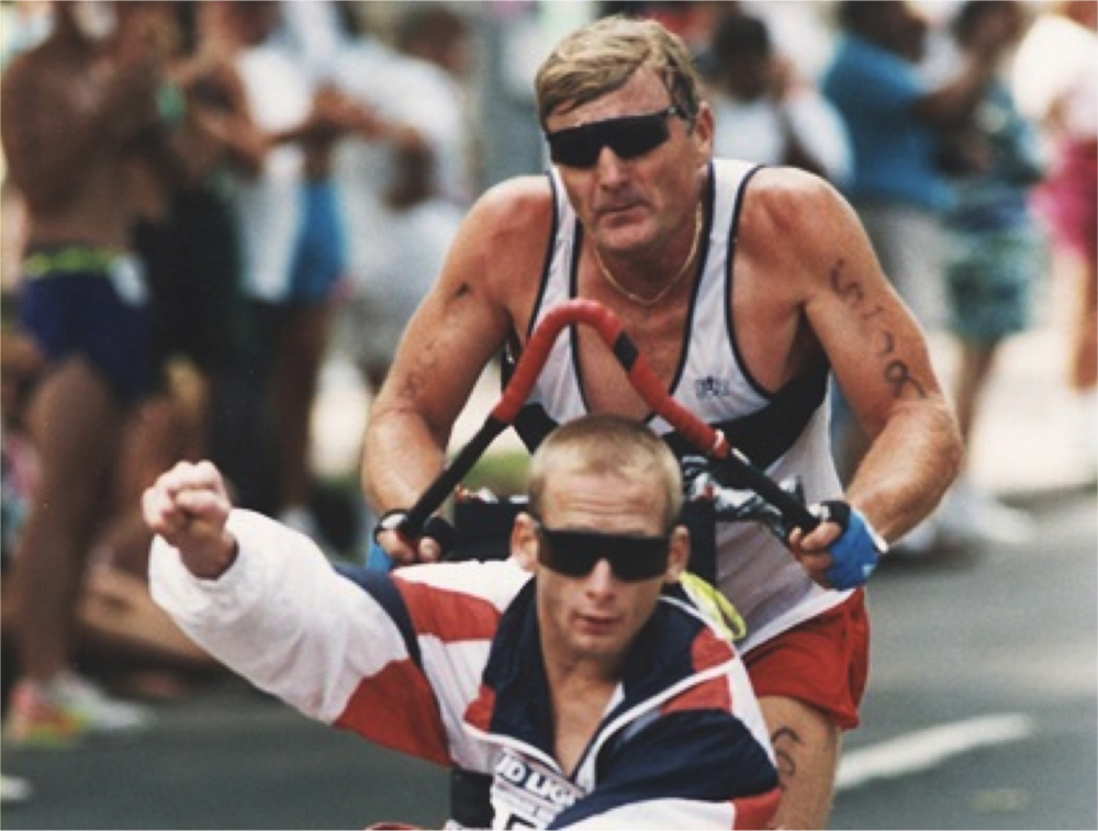 Dick Hoyt pushing his son Rick in a racing chair during an IRONMAN race, Rick raising a fist to the crowd
